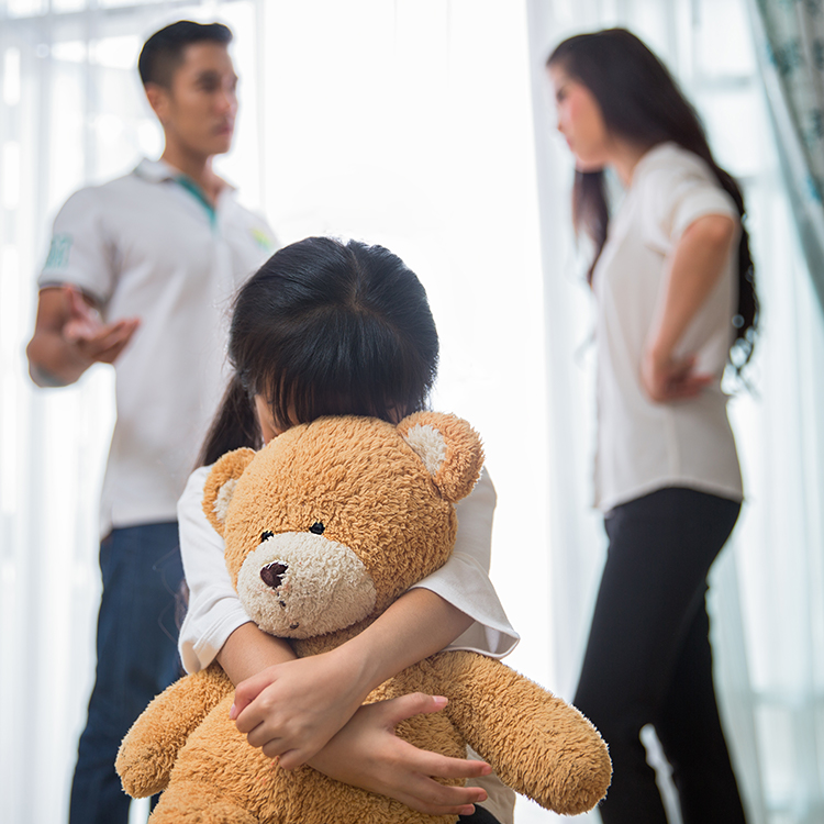 Child holding teddy bear while parents argue in the background
